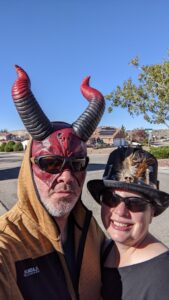 Amanda and Gabe in Halloween get-up. Gabe is wearing a half mask that is a devil mask and has tall curving horns. He has on sunglasses and a orange-ish hoodie. Amanda is wearing a top hat that's made to look like a with doctor hat with feathers and a little skull on it. The day is beautiful, blue sky.