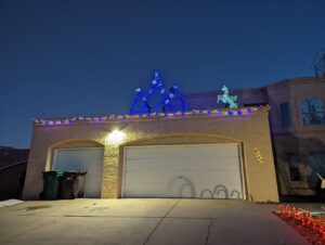 our house. It's a two-story stucco house that looks kind of brown at night, when this photo was taken, and pink during the day. Three car garage. On top is some trees made of wood that are lit up with blue lights and a unicorn-pegasus all lit up. Candy lights are making shadows on the garage.