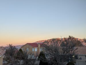 A view from our back deck overlooking the neighborhood and the Sandia Mountains are lit up watermelon color - that's where their name comes from - in the distance. The sky is a perfect sunset of blue fading to orange at the horizon.